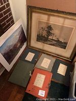 Photo showing various vintage and signed prints, includes framed landscape prints and old portfolios with labels.