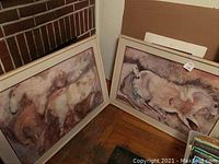 Two horse prints framed, resting against a brick wall with visible artist signature on each print