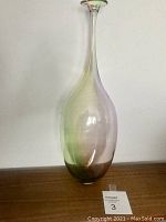 Tall vintage glass vase with slender neck and flared mouth, placed on wood surface against white wall.