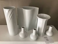 Six white porcelain vases of varying shapes and heights arranged on a white surface against a beige wall.