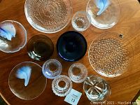 Top-down photo showing a collection of twelve vintage Scandinavian art glass pieces on a wood surface, including clear bowls, plates, and distinctive cobalt blue and textured pieces arranged in a somewhat circular layout.