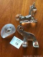 Three clear and frosted glass animal sculptures on wooden surface: an elephant, a unicorn, and a seal with a MaxSold auction number card.