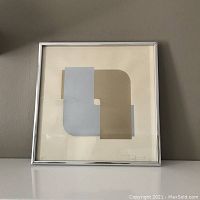 Front view of framed abstract art print showing overlapping gold and silver geometric shapes with a signature visible at lower right.