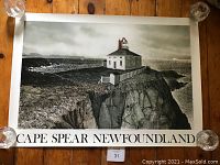 Full view of the Cape Spear Newfoundland print depicting the lighthouse and coastal rocks, resting on a wooden floor held down by glass weights on each corner.