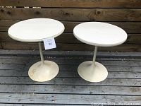 Front angle view of two vintage white metal pedestal side tables showing round tops and bases with surface rust and wear.