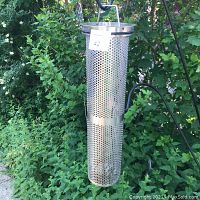Photo of a tall vintage industrial stainless steel light fixture with perforated cylindrical body and conical top shown outdoors with greenery background.