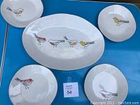 Five ceramic plates displayed on blue surface; one oval serving platter with five birds, four round plates each with one bird.
