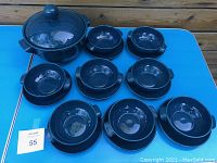 Overall view of large covered dark green ceramic serving tureen, eight matching bowls, and eight plates arranged on blue table.