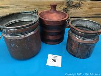 Front view of two handmade copper pots with patinated metal and hand-designed carved handles alongside a vintage wooden container with lid and dark horizontal stripes.