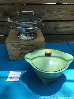 Fire King glass food warmer on stainless steel base with lid and original box behind, green enamel casserole with lid beside it.