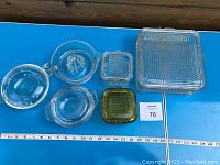 Top-down view of six vintage glass containers for food storage, displaying sizes with a ruler and a large ribbed square container with lid, citrus juicer, lidded round bowl, and smaller square containers in clear and green glass.