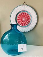 Photo showing the turquoise glass vase in front of the hanging plate with red and white geometric floral design.