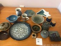 Photo showing a variety of vintage pottery items including plates, bowls, mugs, and jars arranged on a wooden table.
