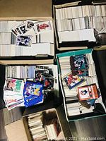 Top view showing four large boxes filled with thousands of trading cards. Cards are stacked vertically and loosely, some spilling out. Variety of sports represented including baseball, hockey, soccer, and basketball. A few cards with player images visible on top.