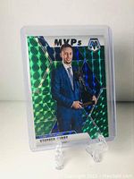 Photo of the Stephen Curry Mosaic MVP green holographic basketball card in a protective plastic holder on a clear display stand
