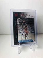 Front side of the 1997 The Big Picture Michael Jordan sample card showing Michael Jordan about to shoot a basketball. Card is inside a protective clear plastic case and displayed on a small clear plastic stand.