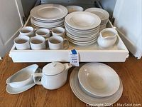 Two sets of white dishes stacked in a cabinet drawer with various plates, bowls, coffee cups, teapot, gravy boat, and serving dishes arranged on a wooden surface.