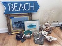 Photo shows the overall contents of the lot including framed artwork, bowls, shells, basket, and the beach sign on the floor.