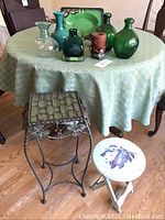 Green glass and pottery vases on a green tablecloth covered round table with metal and wooden plant stands in front