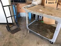Side view of utility rolling work bench with stained surface and metal hand truck beside it.