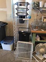 Wide angle showing the full tower of white metal wire organizer with five mesh drawers on wheels alongside plastic bins and a black garbage container with handle.