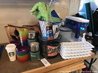 Overview of all lot items on table including thermoses, plastic bowls, canner, ice trays, water jug, and straw tote