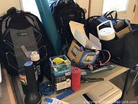 Wide view of lot showing backpacks, blue yoga mat, cooler bags, Coleman thermos, boxed Garmin GPS, and camping accessories on table.