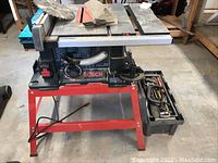 Front view of Bosch table saw mounted on red metal stand with toolbox at right