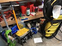 Wide view showing Karcher pressure washer, Ryobi tiller attachment, various garden tools and accessories on floor and shelving.