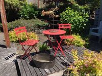 Full view of red metal bistro table with two matching folding chairs, metal lobster wind vane on the table, and metal bucket on deck.