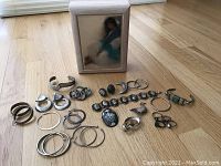 Photo showing wood music box with framed tile art of a woman and various silver jewelry including rings, bracelets, earrings and pins.