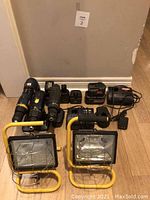 Overall lot arranged on floor showing three cordless drills, multiple chargers and batteries, and two yellow metal-framed portable work lights without bulbs.