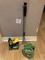 Photo showing new green garden hose with yellow water sprayer in packaging, Miracle-Gro Liquafeed bottle, and silver retractable hand rake leaning against wall on wooden floor.