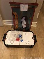 Photo shows the assembled air powered hockey table with white playing surface marked like a hockey rink, with two strikers (one red, one blue) and a puck inside a plastic bag on top. Black handles and black supports are visible.