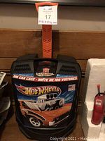 Front view of black plastic Hot Wheels carrying case with flame graphics, showing the 100 car capacity label and retractable handle in orange.