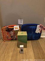 Two Disney lunchboxes, one Toy Story with Woody design in orange/yellow, one Frozen design in dark blue, with price tag showing $12.98 on Frozen lunchbox. Green thermal porcelain cup box and a small white boxed item are placed in front.