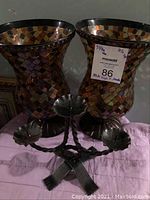 Front view of two mosaic glass hurricane candle holders and the cast iron three-arm candle holder in front of them on a cloth surface, with a MaxSold tag on one glass holder.