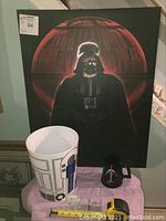 Framed Darth Vader artwork with R2-D2 garbage pail, Darth Vader mug, and two small Star Wars vehicles on a purple cloth
