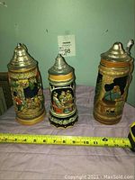 Front view of three beer steins on fabric with tape measure at base showing approximate height of 8-10 inches. Center stein shows colorful musical Moulin Rouge scene, flanked by two traditional scene steins.