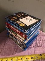 Stack of 12 Blu-ray movies with a measuring tape at bottom showing approximate size and a paper label with number 100 on top.