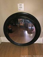 Front view of round black framed mirror with bronze accents, reflecting wooden floor and surroundings. Mirror needs cleaning.