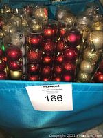 Blue bin filled with multiple tubes/packages of Christmas ball ornaments. Colors include red, gold, green, and silver. Some packages opened with visible wear and scratches on ornaments.