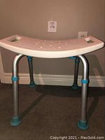 Front view of the adjustable shower seat showing white plastic curved seat with textured surface and metal legs with blue rubber feet.