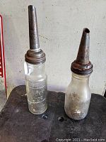 Full view of two antique oil bottles with metal pouring spouts, one clear glass embossed Socony bottle, the other frosted/dirt-covered bottle.