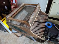 Photo showing the overall wooden cranberry scoop with 16 wooden tines and metal mesh bottom, placed on the floor and surrounded by other items.