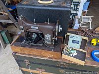 Side view of the sewing machine mounted on a wooden base, showing the machine, case, instruction manual and box of accessories on a wooden surface.