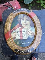Front view of oval sign showing girl holding codfish box, wood-grain border, red band