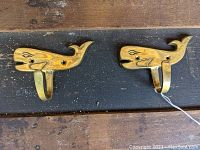 Photo showing two cast brass whale-shaped coat hooks on a wooden surface.