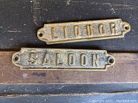 Photo of two bronze plaques, one marked 'LIQUOR' and the other 'SALOON', placed on wood surface.