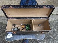 Open view of vintage black tackle box showing fishing reels and fishing lures inside compartments. Base lined with tan material. Tackle box shows wear.
